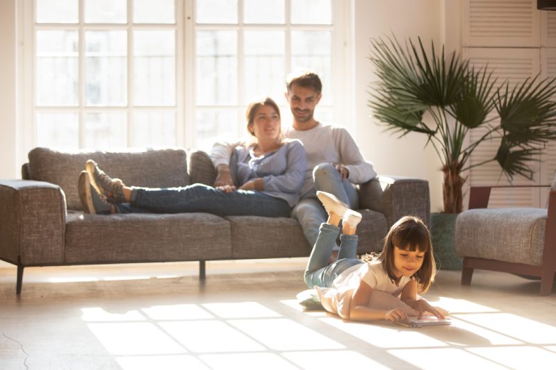 Happy parents relaxing on couch in comfort light living room while little kid child daughter playing on warm floor drawing with colored pencils, family having fun together, underfloor heating concept