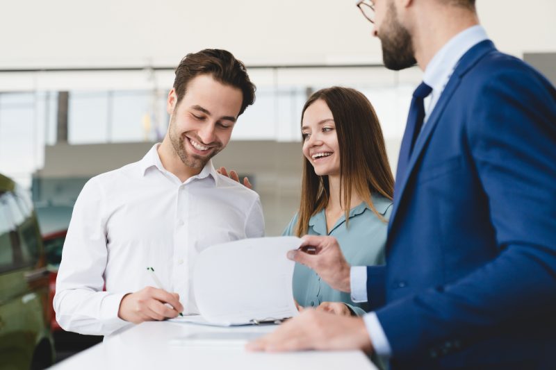 Buying purchasing new car. Male shop assistant congratulating young family couple with buying auto while man signing contract loan mortgage.