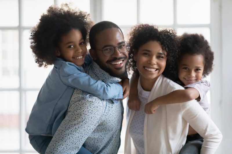 Portrait of African ethnicity parents piggy back son and daughter posing indoors. Pretty family looking at camera photo shoot in modern apartment at home, happy parenthood family services ad concept