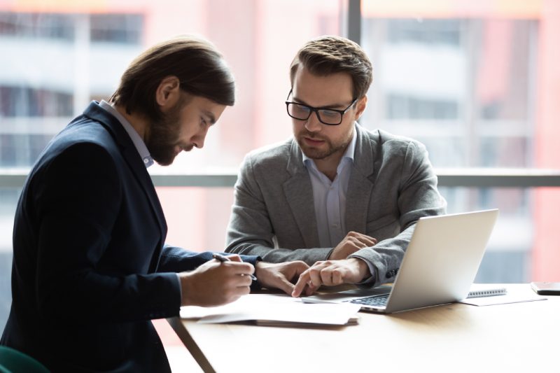 Focused young businessman signing agreement with skilled lawyer in eyeglasses. Concentrated financial advisor showing place for signature on paper contract document to male client at meeting in office