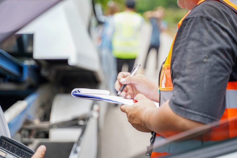Closeup and crop insurance agent writing on clipboard while examining car after accident claim being assessed and processed on blurred damaged car truck slides and group of people background.