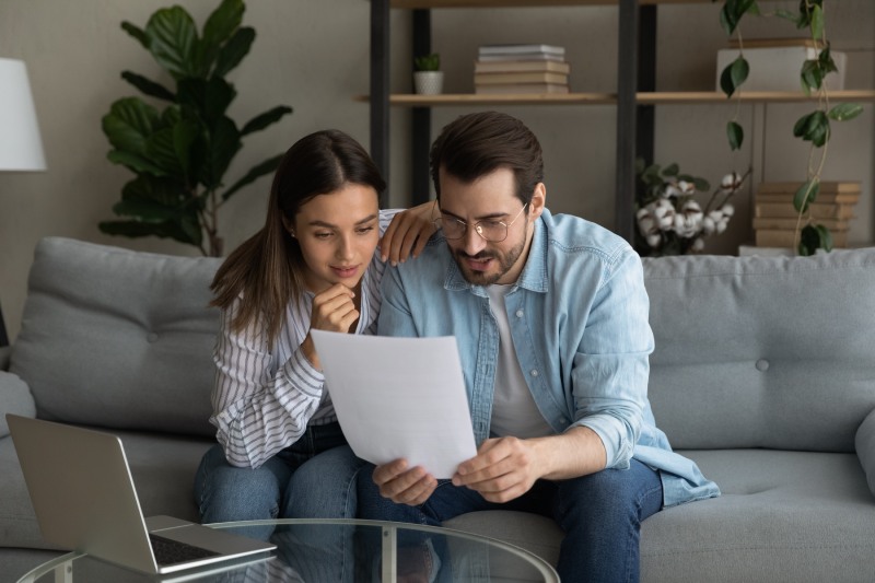 Curious interested young wife and husband couple reading paper document, bank loan approval notification, correspondence letter or considering agreement contract, sitting together on couch at home.