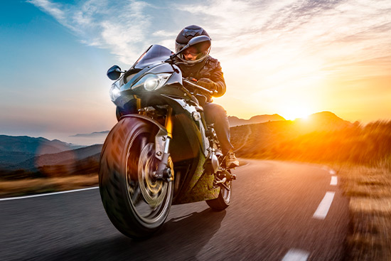 A motorcyclist cruises along a road at sunset, with warm hues illuminating the scene, evoking a sense of exploration.