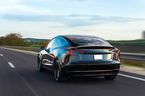 Un coche eléctrico negro circula por una autopista vacía con vegetación y un cielo azul de fondo.