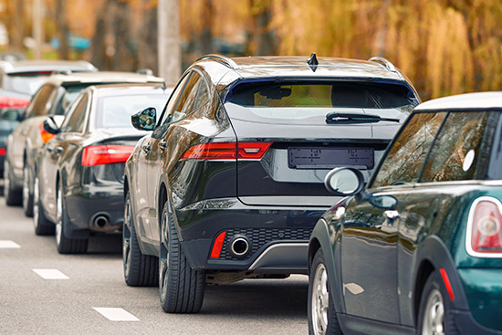 Una fila de coches está aparcada a lo largo de una calle, con árboles otoñales visibles al fondo.